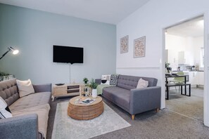 A living room with a grey sofa, a round coffee table, a flat-screen TV mounted on the wall, and a kitchen visible through an open doorway. - Spacious Home Ideal for Contractors Sleeps 7 (Manchester)