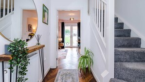 Un pasillo con escalera de madera, un radiador, un espejo, una planta y vistas a una habitación con una puerta.