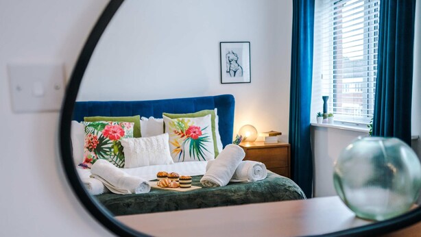 Una cama con almohadones florales, una lámpara de noche y una ventana con cortinas.