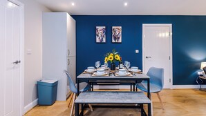 A dining area with a table set for four, two wall-mounted paintings, a trash bin, and a door. - Modern City Centre 3 Bed Apartment Great for Couples (Chester)