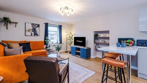 A modern living room with a sofa, coffee table, and TV, a kitchen area with bar stools, and a dining table. - Modern City Centre 3 Bed Apartment Great for Couples (Chester)