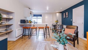A modern kitchen with bar stools, a dining area with a table and chairs, and a living room with a sofa and a decorative plant. - Modern City Centre 3 Bed Apartment Great for Couples (Chester)