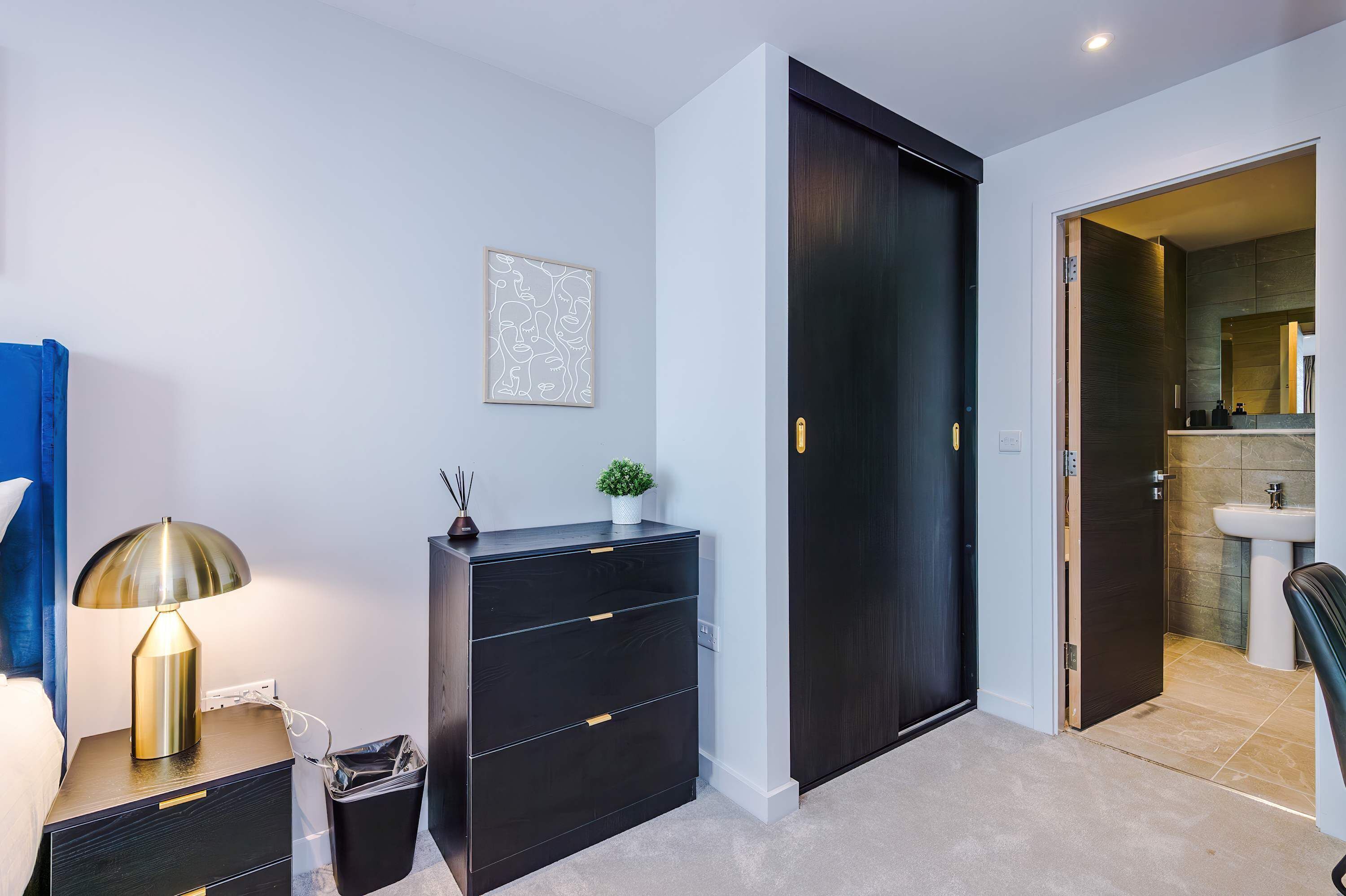 A modern room with a bed, bedside lamp, black dresser, and a door leading to a bathroom.