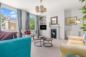 A living room with a sofa, a round table, a television, a fireplace, and a window with curtains. - Chic 2BR Duplex Home W London With Private Patio (London)