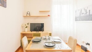 Ein Essbereich mit einem Tisch für vier Personen, einem Fernseher auf einem Standfuß und einem Fenster mit durchscheinenden Vorhängen.