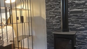 Une pièce comprenant un lit, une étagère en métal noir, un mur en pierre et un poêle à bois.