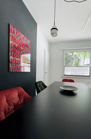 Une pièce moderne avec une table noire, des chaises rouges et vertes, un grand tableau abstrait et une fenêtre donnant sur des arbres.