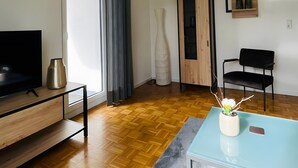 Una sala de estar moderna con piso de madera, una mesa de centro de vidrio, un televisor sobre un mueble de madera, una silla negra, un jarrón blanco con flores y un mueble de madera.