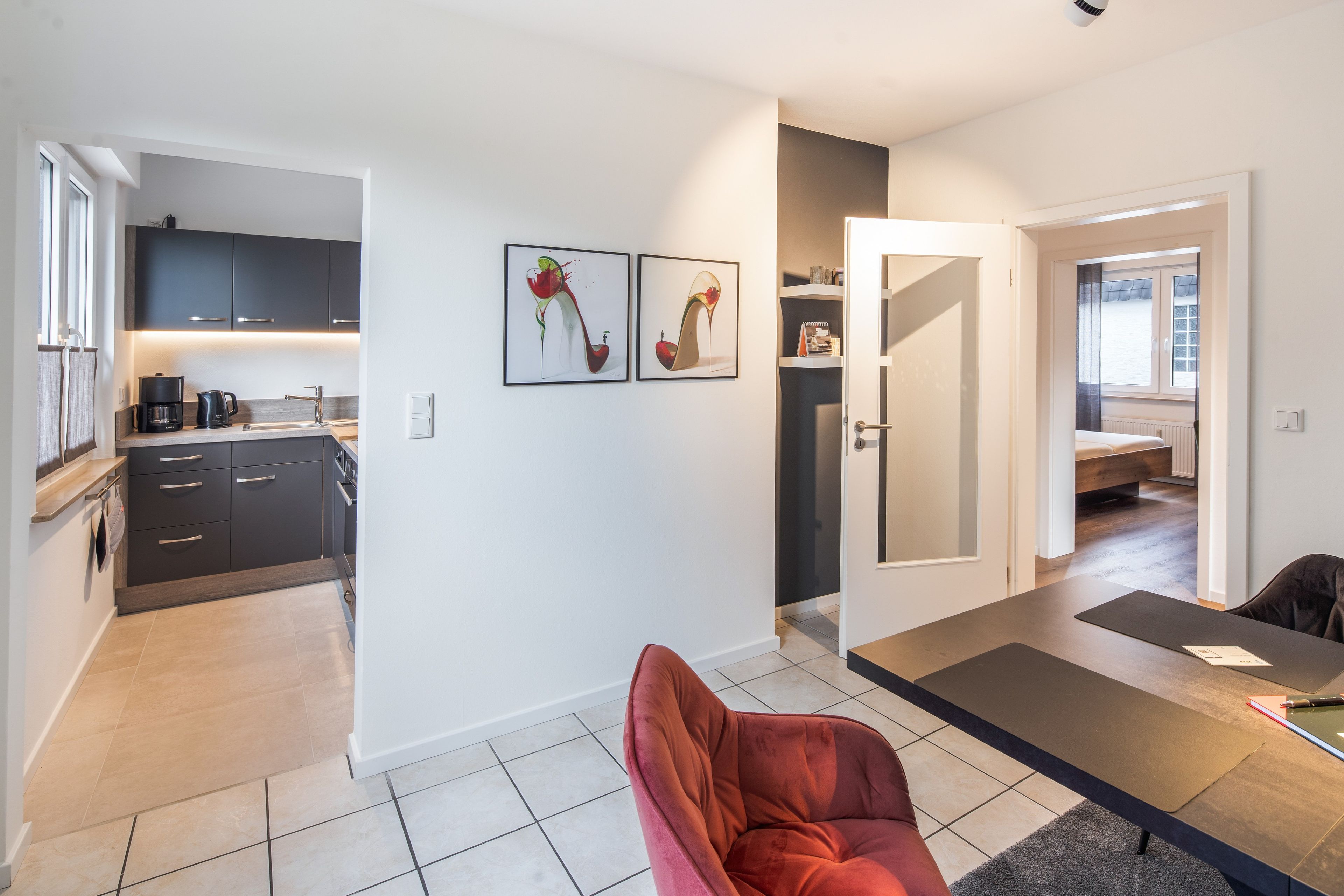 A modern kitchen with dark cabinets, a coffee machine, and a dining area with a red chair and a table.