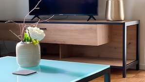 Una sala de estar moderna con un televisor de pantalla plana, un escritorio de madera, una mesa de centro azul claro, un jarrón blanco con flores y un teléfono inteligente.