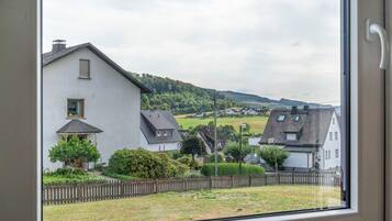 Blick durch ein Fenster auf eine Wohngegend mit Häusern, einem Holzzaun und einer Hügelkulisse im Hintergrund.