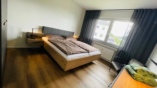 Une chambre à coucher avec un lit en bois, une table de chevet, une lampe, une fenêtre avec des rideaux et une vue sur les bâtiments à l’extérieur.