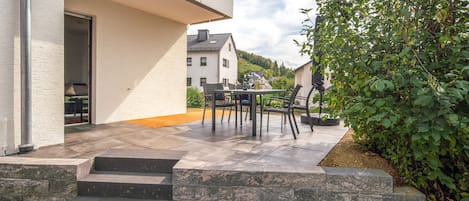 A patio with outdoor furniture and a table, steps leading to a building entrance, and a garden area with a hedge.
