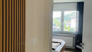 Un dormitorio con cama, radiador, ventana que ofrece vistas a casas y vegetación, y puerta con detalles de panel de madera.