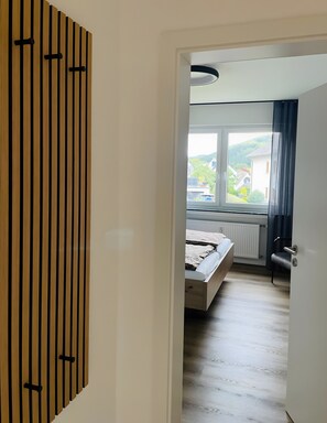 Un dormitorio con cama, radiador, ventana que ofrece vistas a casas y vegetación, y puerta con detalles de panel de madera.