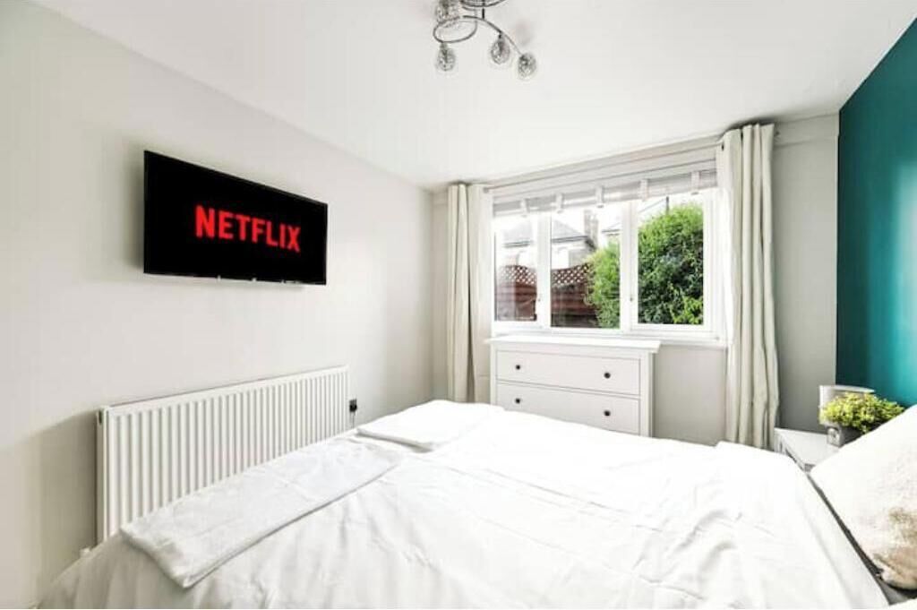 Een slaapkamer met een bed, een witte dressoir, een raam met gordijnen, een radiator en een televisie aan de muur waarop 'NETFLIX' wordt weergegeven.