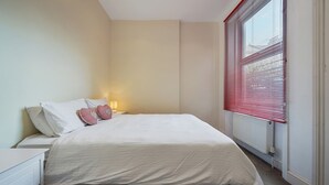 Een slaapkamer met een bed, nachtkastje, lamp en raam met jaloezieën.