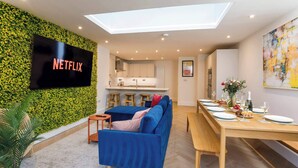 Ett modernt vardagsrum med en platt-TV som visar "NETFLIX", en matplats med ett träbord, en blå soffa och ett kök i bakgrunden.