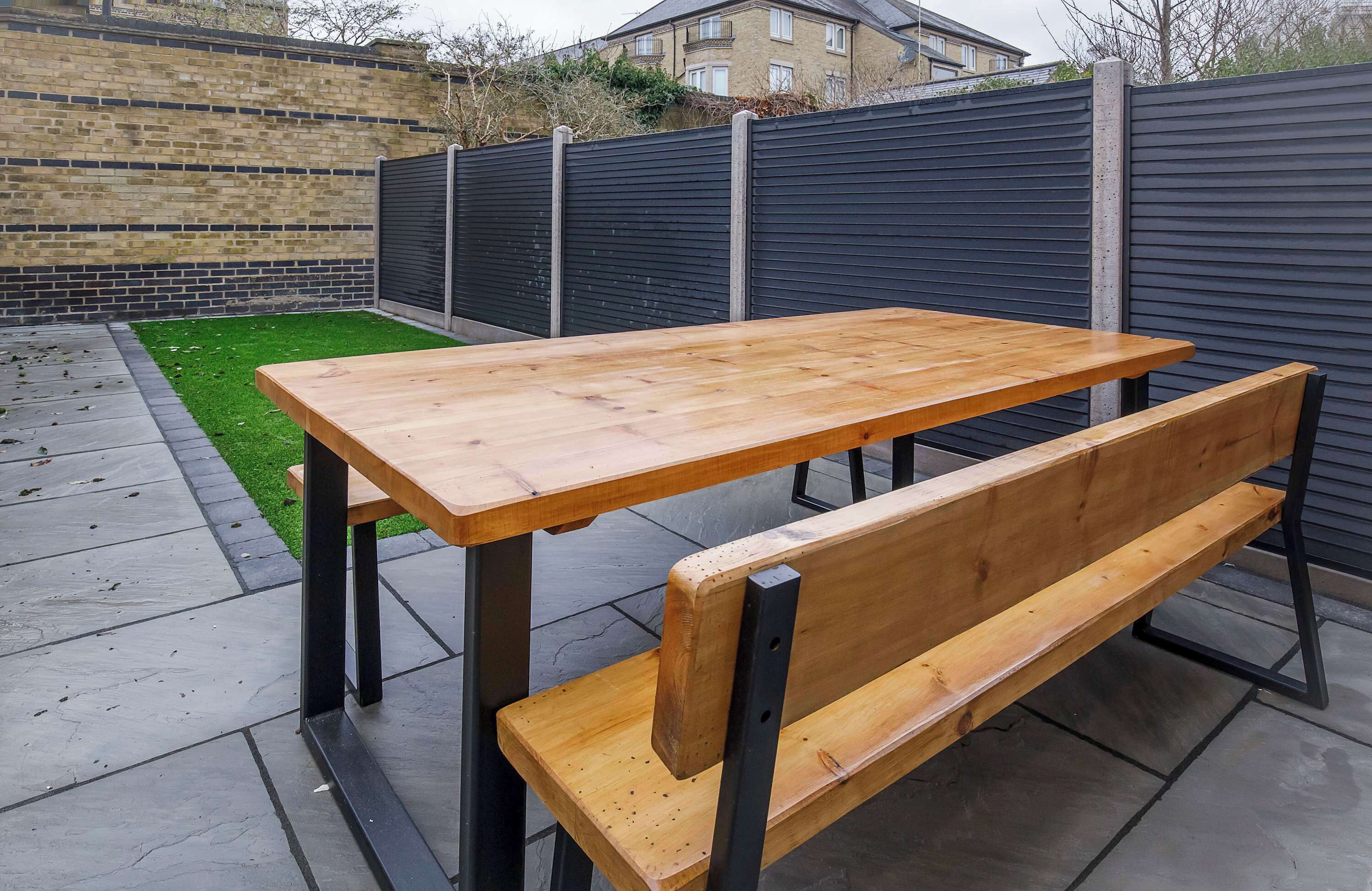 A wooden table and bench set outdoors, with a brick wall and a black fence in the background.