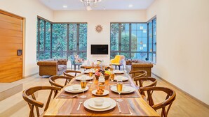 A dining room with a wooden table set for six, surrounded by wooden chairs, a chandelier, and large windows with a view of greenery. - ELIVAAS Villa 39 Serene 6BHK with Pool (Lonavala)