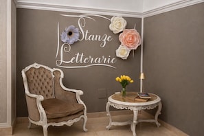 A room with a tufted armchair, a small round table with a book and flowers, and a wall with a decorative sign. - Hotiday Catania Stanze Letterarie (Catania)