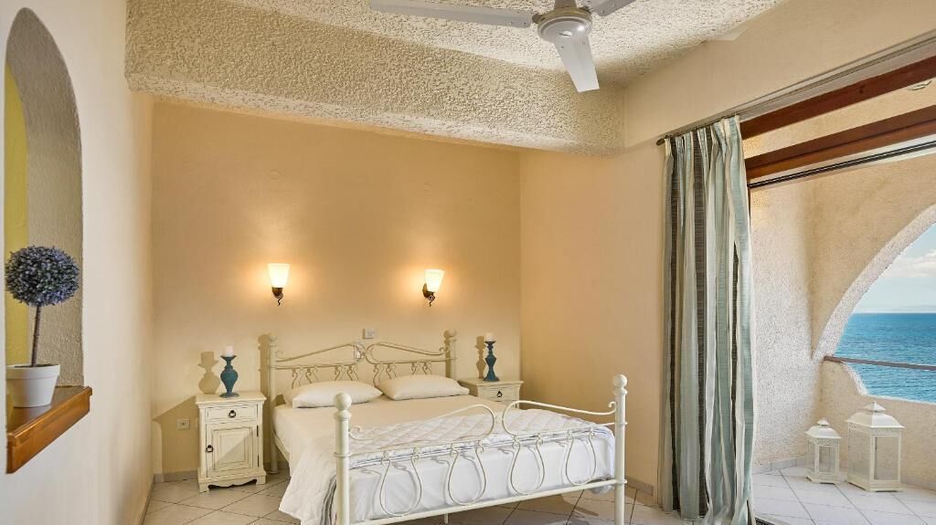A bedroom with a bed, bedside tables, wall sconces, a ceiling fan, and a view of the sea.