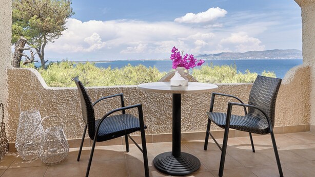 Eine Terrasse mit Tisch, zwei Stühlen, einer Blumenvase und Blick auf Meer und Hügel.