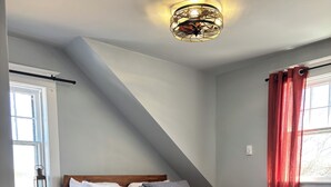 A bedroom with a sloped ceiling, a bed with grey bedding, a wooden dresser, and a window with red curtains. - Coastal Trail Cottages - Captain Jack's Lookout (Morell)