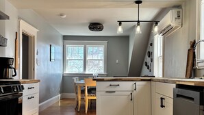 A modern kitchen with white cabinetry, a built-in oven, and a dishwasher. There is a dining area with a table and chairs, and a window providing natural light. - Coastal Trail Cottages - Captain Jack's Lookout (Morell)