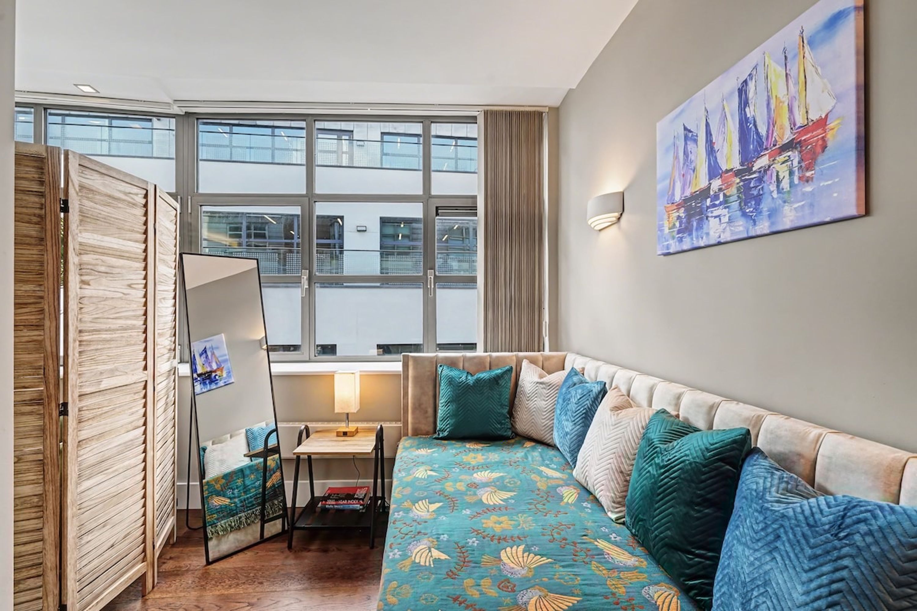 A room with a large window, a bed with patterned bedding, a desk with a lamp, and a painting on the wall.