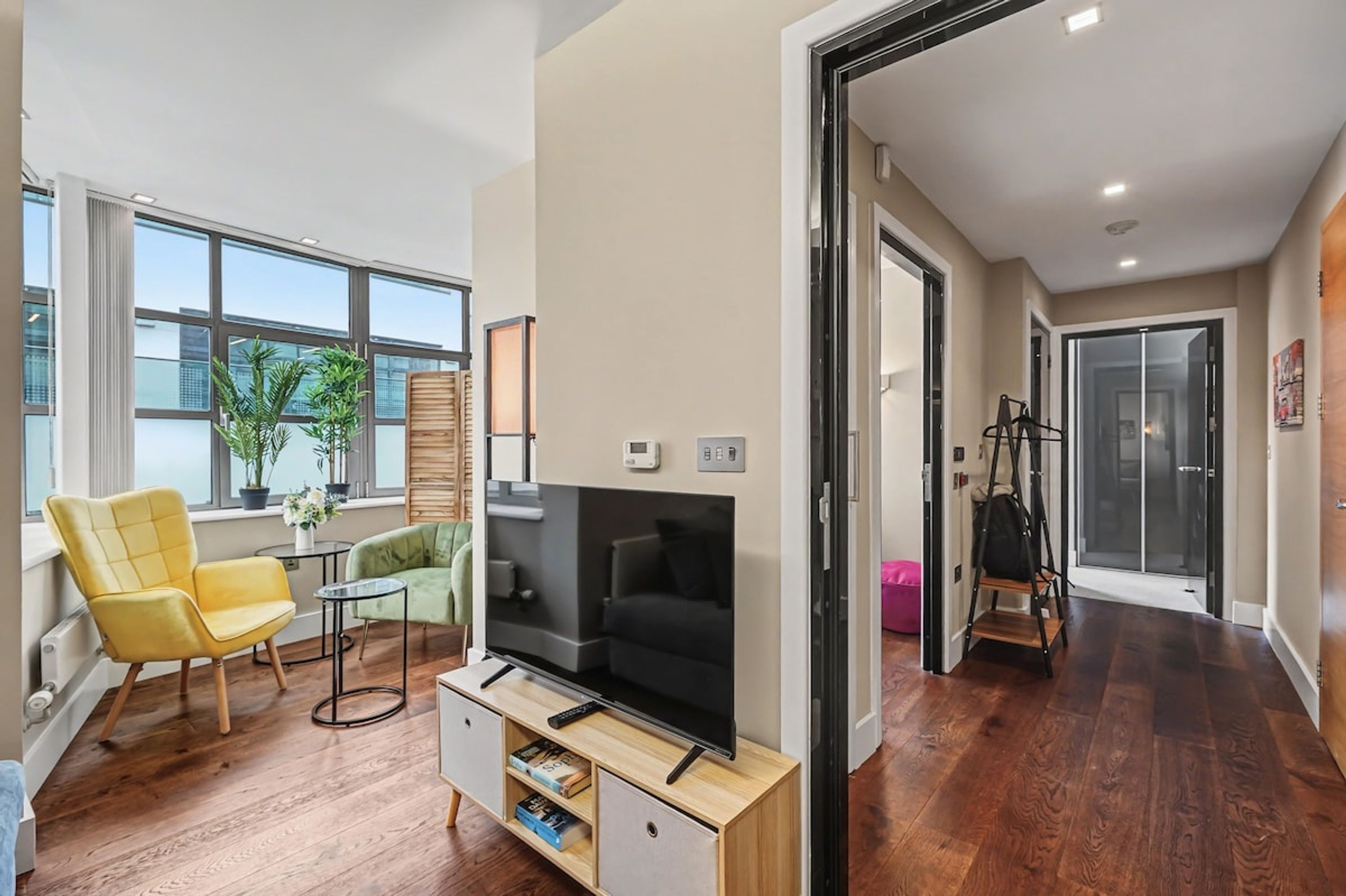 Una sala de estar moderna con un televisor de pantalla plana, piso de madera, un sillón amarillo, un sofá verde y una pequeña mesa con una planta en maceta.