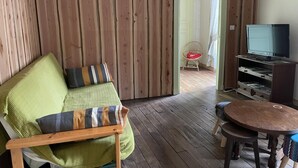 Une pièce avec un plancher en bois, un canapé vert avec des coussins à rayures, une chaise en bois, une petite table en bois et un téléviseur posé sur un meuble en bois.