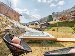Een hottub op een houten terras, met stenen muren, omringd door tuinmeubels, met op de achtergrond huizen en een glooiend landschap.