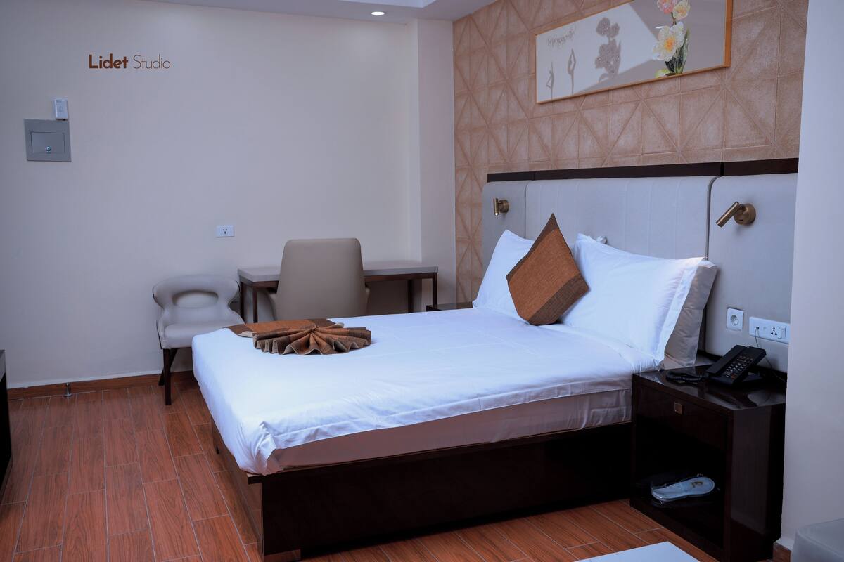 A hotel room with a bed, a desk, a chair, a nightstand, and a wall-mounted artwork.