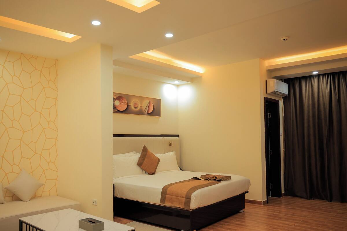 A hotel room with a bed, a sofa, a painting, and a ceiling light.