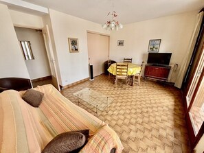 A living room with a sofa, a dining table with chairs, a television, and a fireplace. - Bright apartment, terrace, private parking (Le Lavandou)
