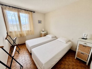 A room with two beds, a chair, a small table, a radiator, and a window with curtains. - Bright apartment, terrace, private parking (Le Lavandou)