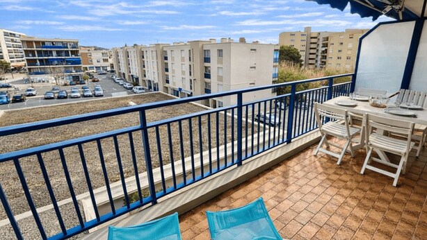 Een balkon met uitzicht op een parkeerplaats en gebouwen, een eettafel en blauwe stoelen.