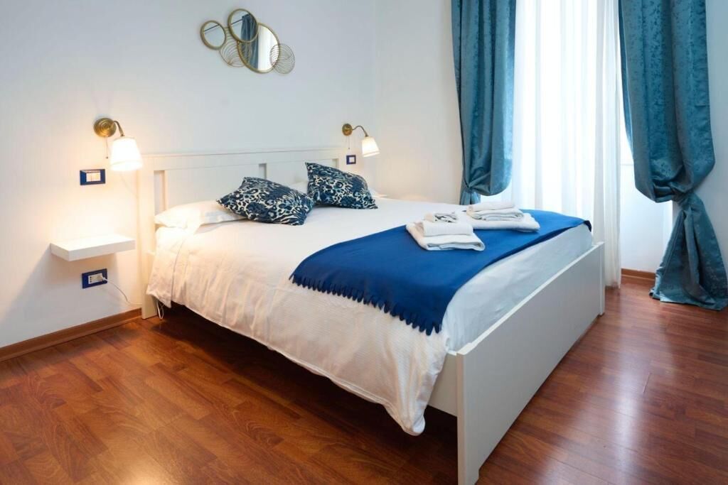 A neatly made bed with a blue and white bedspread, two bedside lamps, a mirror above the bed, and a window with curtains.
