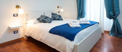 A neatly made bed with a blue and white bedspread, two bedside lamps, a mirror above the bed, and a window with curtains.
