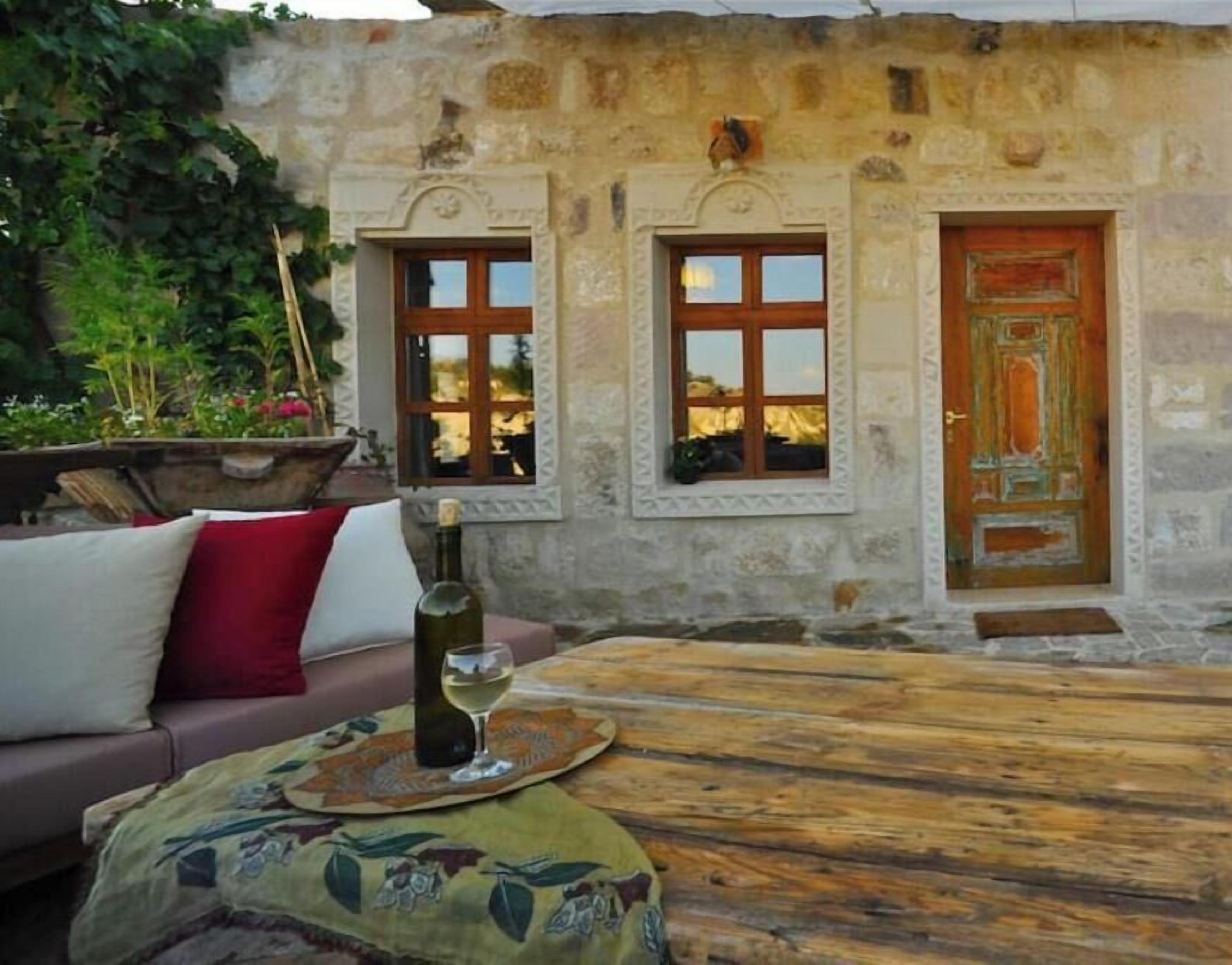 Un edificio rústico de piedra con dos ventanas y una puerta de madera, una mesa de madera con una botella y un vaso, y un sofá con cojines.