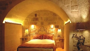 Quarto com teto em arco de pedra, uma cama com cabeceira, duas luminárias de apoio e um abajur fixado na parede.