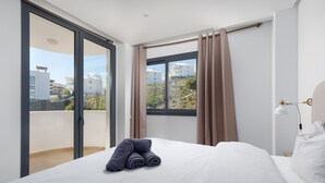 Een hotelkamer met een bed, een raam met gordijnen, een balkon en uitzicht op gebouwen.