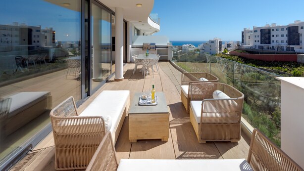 Een balkon met rieten meubilair, een tafel met een tijdschrift, en een glazen hekwerk met uitzicht op een stadsgezicht.