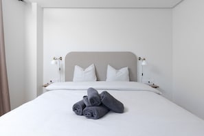 En pænt redt seng med hvide sengetæpper og sammenrullet håndklæder, to sengeborde med lamper, og en enkel sengegavl.