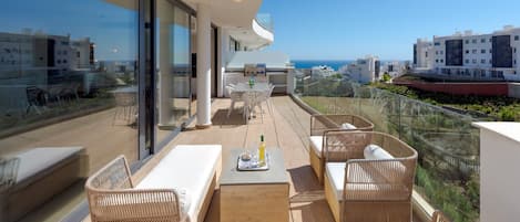 A balcony with wicker furniture, a wooden coffee table, and a glass railing overlooking a coastal cityscape.