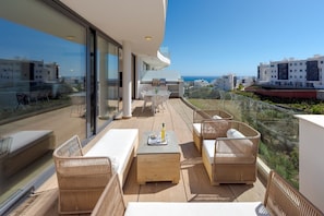 Ein Balkon mit Korbmöbeln, einem Holztisch und einem Glasgeländer mit Blick auf eine Küstenstadt.