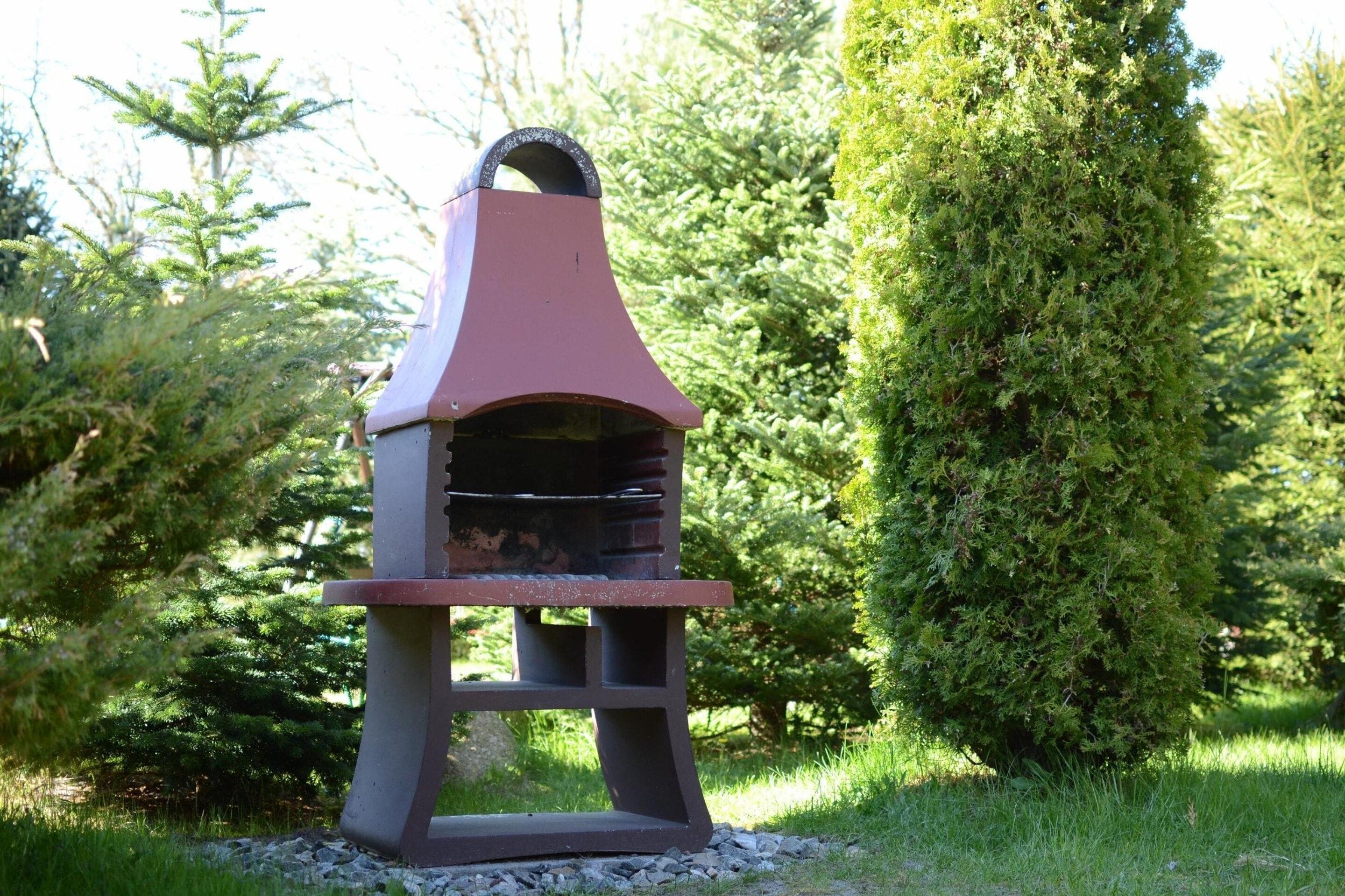 A barbecue grill with a chimney, set in a garden with greenery and trees.
