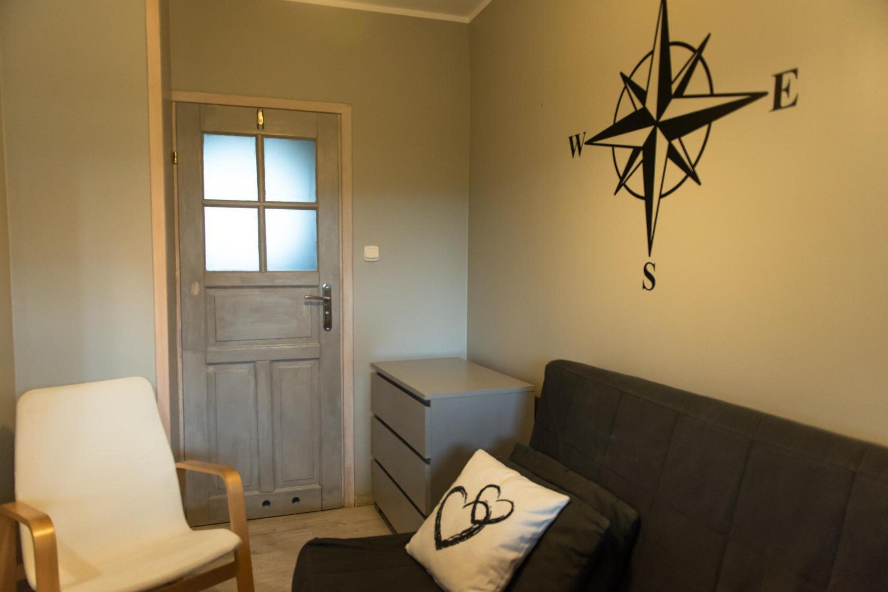 Una habitación con puerta, una silla blanca, un sofá, un pequeño armario blanco y decoración de pared con una brújula.