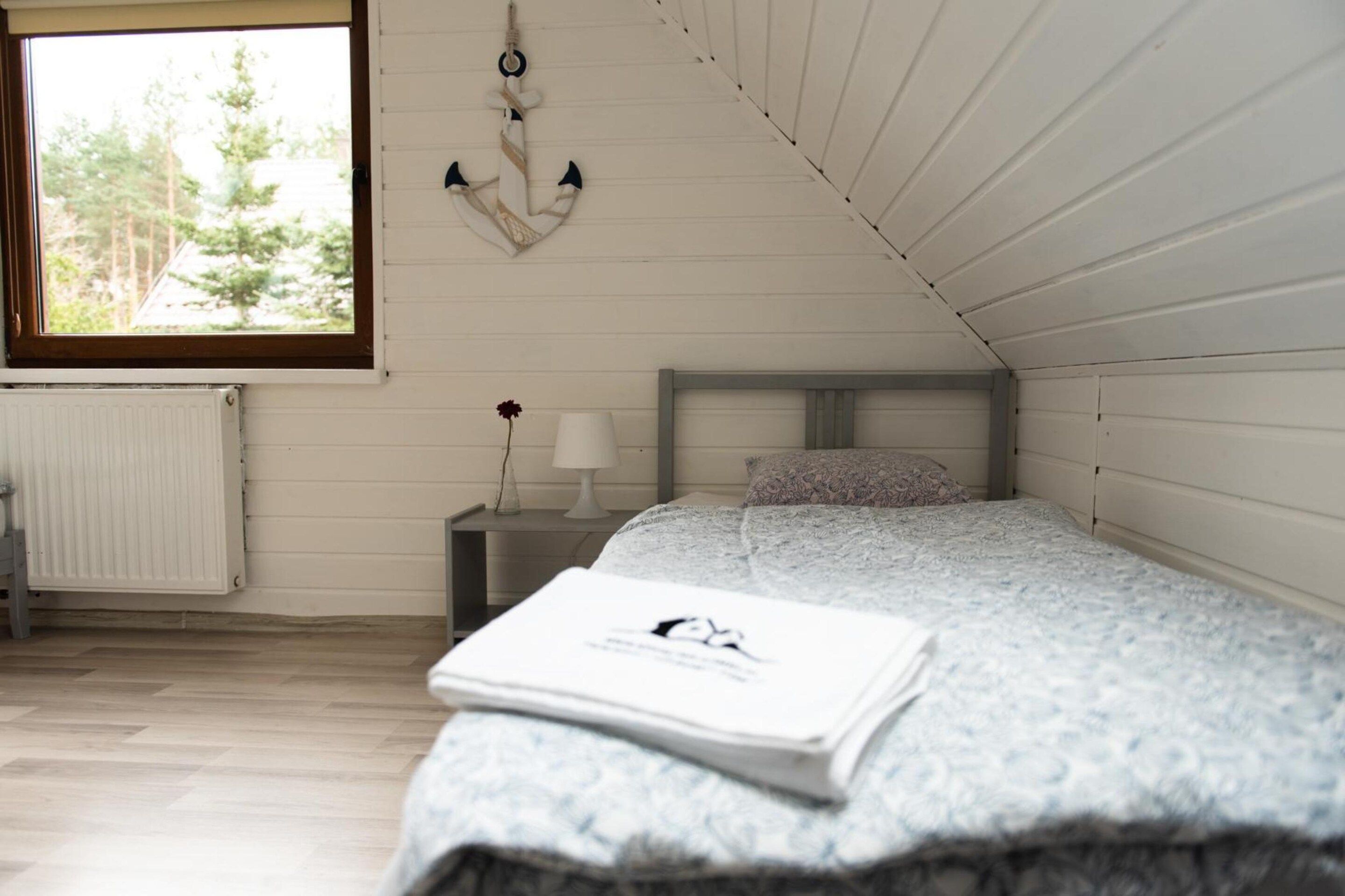 A bedroom with a sloped ceiling, a bed with a patterned bedspread, a bedside table with a lamp, and a window with a view of trees.